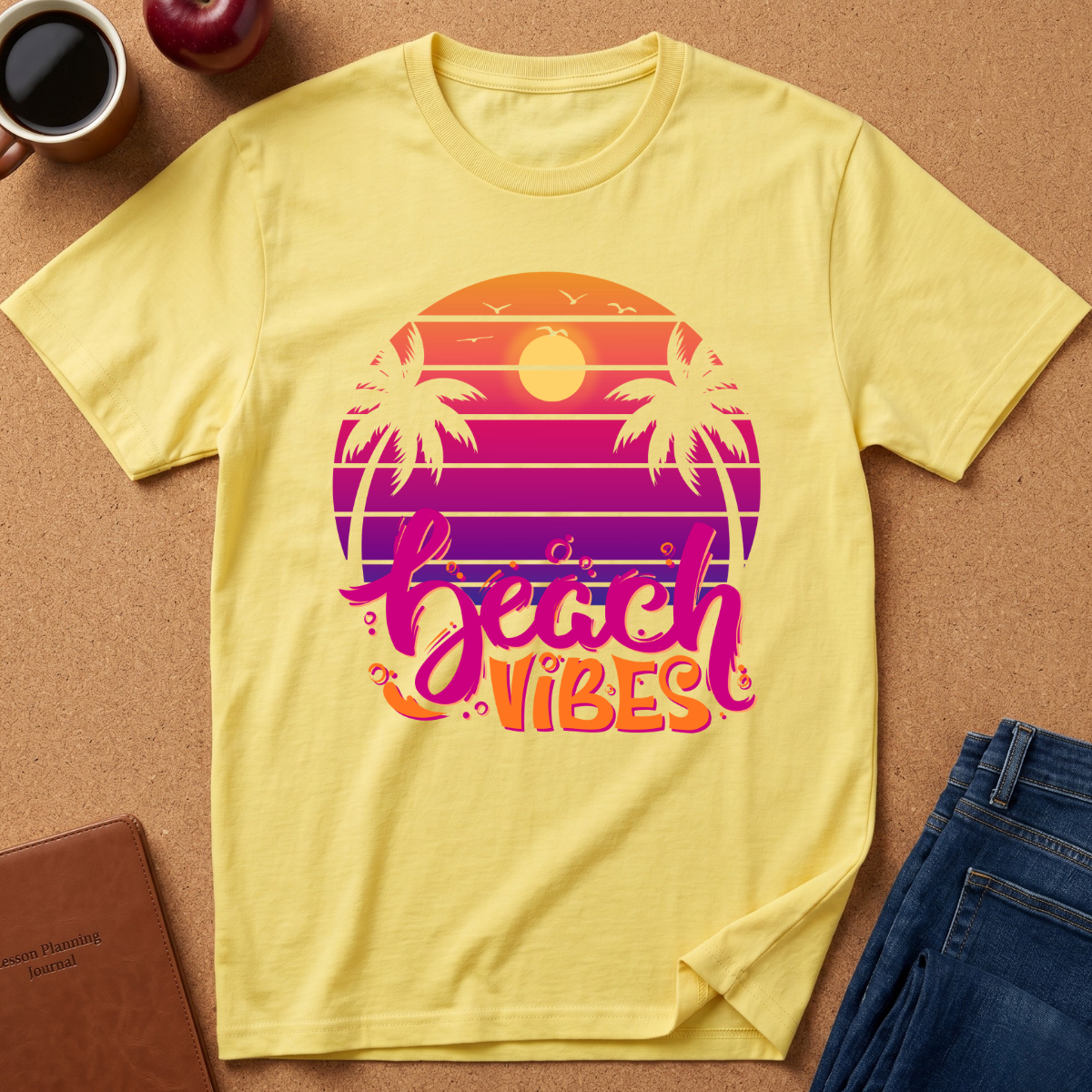 A yellow short-sleeve t-shirt laid flat on a cork surface, featuring a retro sunset graphic with palm trees and the words 'beach vibes' in pink, purple, and orange. A coffee cup, apple, notebook, and jeans are styled around it as props.