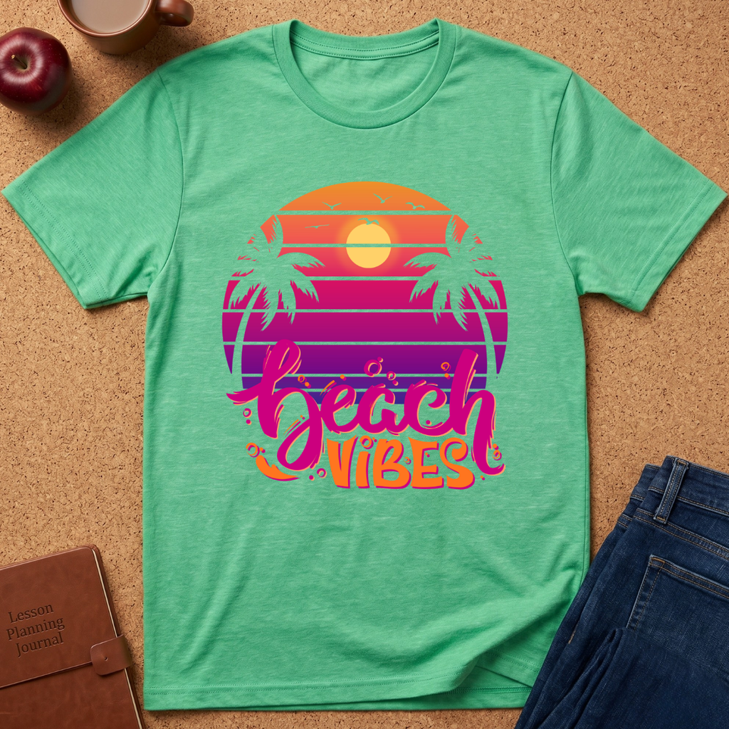 A mint green heather t-shirt laid flat on a cork background, featuring a retro sunset graphic with palm trees and the text 'beach vibes' in pink and orange. Styled flat-lay props include a brown leather journal, a red apple, a coffee mug, and a pair of blue jeans.