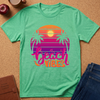 A mint green heather t-shirt laid flat on a cork background, featuring a retro sunset graphic with palm trees and the text 'beach vibes' in pink and orange. Styled flat-lay props include a brown leather journal, a red apple, a coffee mug, and a pair of blue jeans.