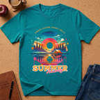 A teal short-sleeve graphic T-shirt laid flat on a cork surface, surrounded by lifestyle props including a coffee cup, apple, notebook, pen, and blue jeans. The shirt features a colorful retro mountain sunset campsite print with the words 'Summer Freedom Journey Again' and 'Summer Journey'.