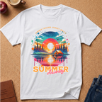 A white short-sleeve t-shirt with a colorful retro summer camping graphic, laid flat on a cork background with lifestyle props like an apple, a coffee cup, and jeans.