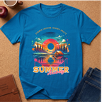 A bright blue short-sleeve t-shirt laid flat on a cork surface, featuring a colorful retro-style summer travel graphic with a sunset, mountains, trees, tent, and the words 'SUMMER FREEDOM JOURNEY AWAY'. Surrounding props include a coffee cup, apple, brown journal, and blue jeans.