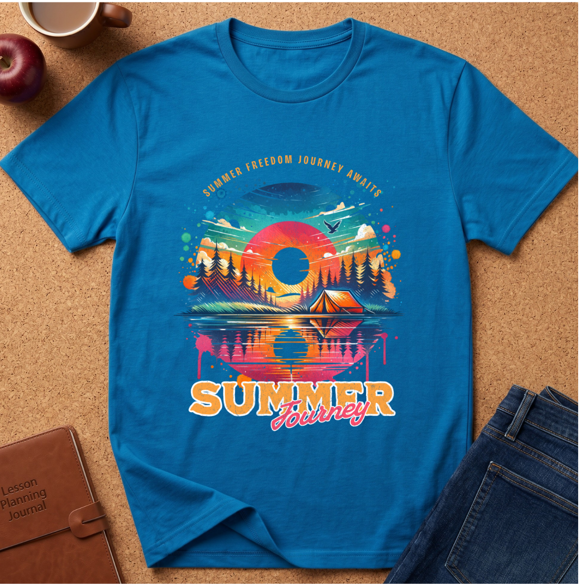 A bright blue short-sleeve t-shirt laid flat on a cork surface, featuring a colorful retro-style summer travel graphic with a sunset, mountains, trees, tent, and the words 'SUMMER FREEDOM JOURNEY AWAY'. Surrounding props include a coffee cup, apple, brown journal, and blue jeans.