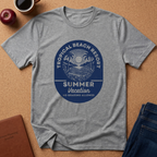 A heather gray short-sleeve t-shirt with a blue circular graphic reading 'Tropical Beach Resort Summer Vacation No Grading Allowed', laid flat on a cork background with a coffee cup, apple, notebook, and jeans nearby.