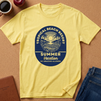 A pale yellow short-sleeve T-shirt laid flat on a tan cork-like surface, featuring a large circular tropical beach resort graphic in dark blue and cream that reads 'Tropical Beach Resort Summer Vacation No Grading Allowed.' Surrounding props include a coffee cup, apple, notebook, and folded jeans.