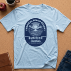 A light blue crewneck t-shirt laid flat on a cork board background, featuring a dark blue circular graphic with palm trees, a sun, and the text 'TROPICAL BEACH RESORT SUMMER Vacation NO GRADING ALLOWED'. A brown leather notebook, a red apple, a coffee mug, and blue jeans are styled around it.