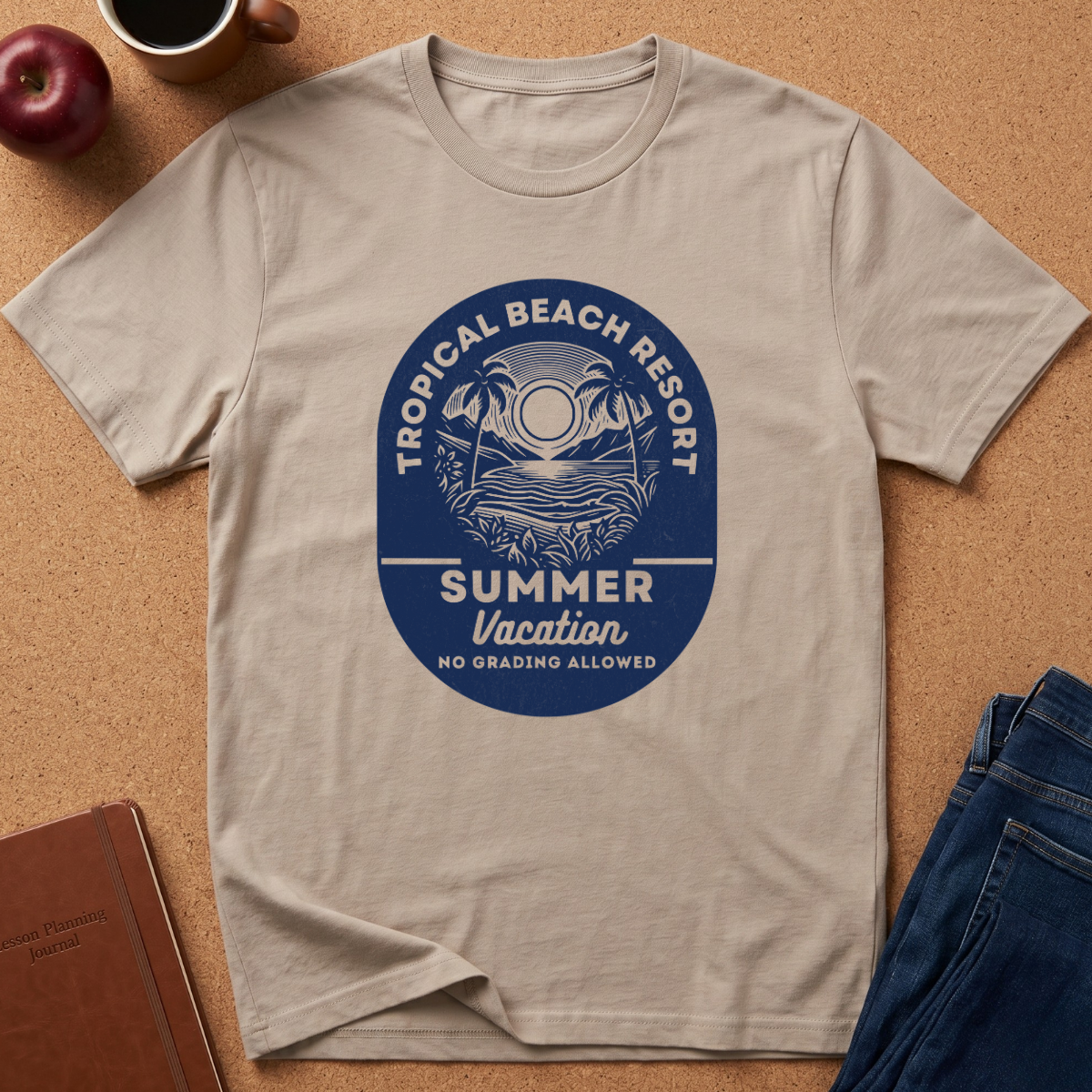 A light beige crewneck t-shirt laid flat on a cork background, featuring a blue circular graphic that reads 'TROPICAL BEACH REPORT SUMMER Vacation NO GRADING ALLOWED' with a palm tree and sun illustration. Props like an apple, coffee cup, notebook, and jeans are visible around it.