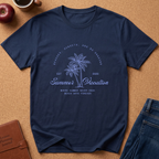 A navy blue short-sleeve t-shirt with a light blue graphic print featuring palm trees and the text 'Summer Vacation' laid flat on a cork background with lifestyle props including jeans, a coffee mug, an apple, and a leather journal.