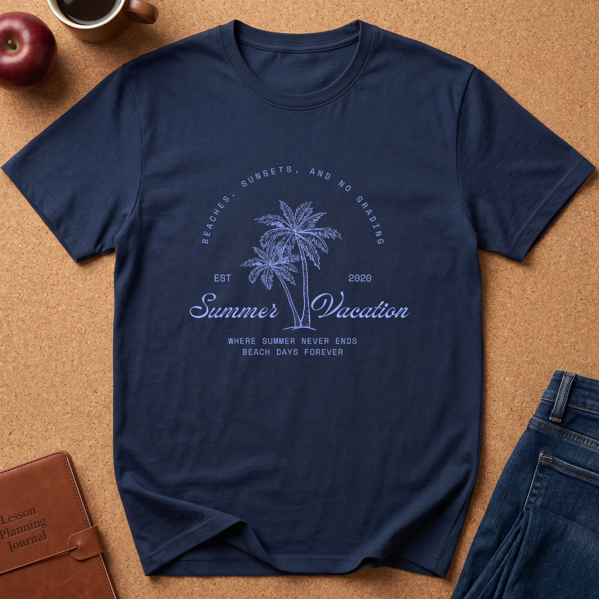 A navy blue short-sleeve t-shirt with a light blue graphic print featuring palm trees and the text 'Summer Vacation' laid flat on a cork background with lifestyle props including jeans, a coffee mug, an apple, and a leather journal.