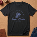 A black short-sleeve graphic t-shirt laid flat on a cork background, featuring a blue palm tree design with the text 'Summer Vacation' and beach-themed phrases. Styled flat-lay props include an apple, a coffee mug, a journal, and blue jeans.