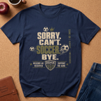 A navy blue graphic t-shirt laid flat on a brown cork background, surrounded by lifestyle props including an apple, a coffee cup, a leather journal, and blue jeans. The shirt features a distressed graphic with the text 'SORRY CAN'T SOCCER BYE.