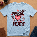 A light blue graphic t-shirt laid flat on a corkboard background, featuring the text 'SCHOOL NURSE A WORK OF HEART' with a pink heart, stethoscope, and medical cross design. Props like an apple, coffee mug, notebook, and jeans are visible around the shirt.