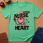 A mint green graphic t-shirt laid flat on a cork background, featuring the text 'SCHOOL NURSE' and 'A HEART IS A NURSE' with a pink heart, stethoscope, and medical cross design. Styled flat-lay props include an apple, coffee cup, book, and jeans.