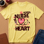A yellow short-sleeve graphic T-shirt laid flat on a cork surface, with a school nurse theme and the phrase 'A WORK OF HEART' in dark brown lettering, pink hearts, and medical icons. Surrounding props include a coffee cup, apple, notebook, and folded jeans.