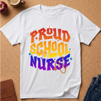 A white short-sleeve T-shirt laid flat on a cork surface, featuring a colorful retro-style graphic that reads “PROUD SCHOOL NURSE” in orange, yellow, and purple lettering, with a stethoscope and nurse cap illustration. Partial props including an apple, coffee cup, book, and jeans are visible around the shirt.