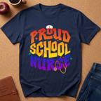 A navy blue graphic t-shirt laid flat on a brown cork board background, featuring the text 'PROUD SCHOOL NURSE' in a colorful retro gradient with a nurse hat and stethoscope illustration. Styled flat-lay props include an apple, a coffee cup, a brown journal, and blue jeans.