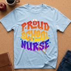 A light blue short-sleeve t-shirt laid flat on a cork background, featuring a colorful retro-style graphic that reads 'PROUD SCHOOL NURSE' in orange, yellow, and purple lettering with a stethoscope and nurse hat illustration. Styled flat-lay props include an apple, coffee mug, notebook, and jeans.