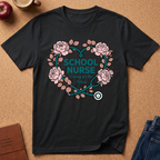 A black short-sleeve graphic T-shirt laid flat on a cork-like surface, featuring a floral heart design with pink roses, teal leaves and stethoscope elements, and the words 'SCHOOL NURSE' and 'Caring for the Future.' Partial props including an apple, mug, journal, and jeans are visible around the shirt.