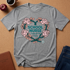 A heather gray short-sleeve T-shirt laid flat on a cork surface, featuring a floral heart graphic with pink roses, a teal stethoscope, and the words 'SCHOOL NURSE' and 'Caring for the Future.' Surrounding props include an apple, a coffee cup, a brown journal, and folded blue jeans.