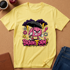 A yellow short-sleeve graphic T-shirt laid flat on a cork-like surface, featuring a cartoon angry pink brain holding a black umbrella with lightning bolts, gears, and the text 'BRAINSTORM!' in pink and yellow. A coffee cup, apple, notebook, and jeans are partially visible around it as styling props.