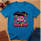 A bright blue short-sleeve graphic T-shirt laid flat on a cork surface, featuring a cartoon angry pink brain holding a black umbrella with lightning bolts, gears, and the text 'BRAINSTORM!' in pink and yellow. Surrounding props include an apple, a coffee mug, a brown journal, and folded blue jeans.