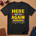 A black short-sleeve graphic T-shirt laid flat on a cork-like surface, featuring the phrase “HERE WE GO AGAIN” in yellow polka-dot letters with red apples, a yellow pencil, and multicolored stars. The shirt is styled with partial jeans, a book, and a cup around it.