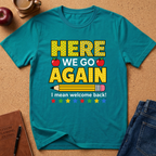 A teal short-sleeve t-shirt laid flat on a cork surface, featuring a school-themed graphic that reads 'HERE WE GO AGAIN I mean welcome back!' with red apples, a pencil, and colorful stars. A brown journal, pen, apple, and blue jeans are partially visible around it as styling props.
