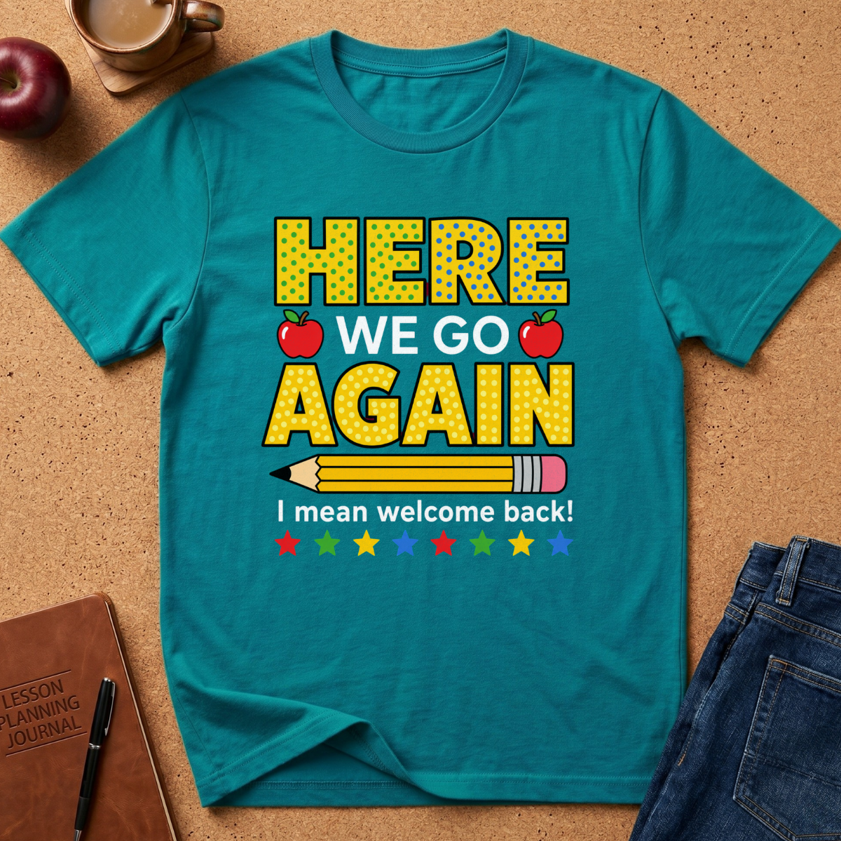 A teal short-sleeve t-shirt laid flat on a cork surface, featuring a school-themed graphic that reads 'HERE WE GO AGAIN I mean welcome back!' with red apples, a pencil, and colorful stars. A brown journal, pen, apple, and blue jeans are partially visible around it as styling props.