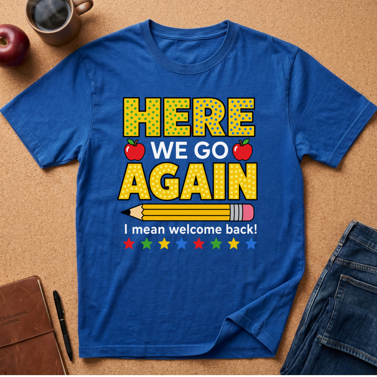 A royal blue graphic t-shirt laid flat on a cork background, featuring a school-themed 'Here We Go Again' print with red apples, a pencil, and colorful stars. Styled flat-lay props include a coffee mug, an apple, a pen, a brown notebook, and folded jeans.