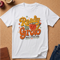 A white short-sleeve graphic T-shirt laid flat on a cork surface, featuring a colorful retro-style 'Ready to Grow Back to School 2026' design with an apple, pencil, and stars. Partial props including an apple, coffee cup, journal, and jeans are visible around the shirt.