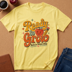 A light yellow graphic t-shirt laid flat on a cork background, featuring a retro-style 'Ready to Grow Back to School 2026' design with an apple and pencil. Styled flat-lay props include a coffee mug, an apple, a notebook, and blue jeans.