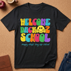 A black graphic t-shirt laid flat on a cork background, featuring a colorful retro-style 'WELCOME BACK 2 SCHOOL' design with a yellow smiley face and the text 'Happy First Day Of School' underneath. Styled flat-lay props include an apple, a coffee cup, a journal, and jeans.
