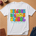 A white short-sleeve T-shirt laid flat on a cork surface, printed with colorful retro-style text reading 'WELCOME BACK 2 SCHOOL' and a yellow smiley face, plus smaller teal script text below. Partial props around the shirt include an apple, a mug, a notebook, and jeans.