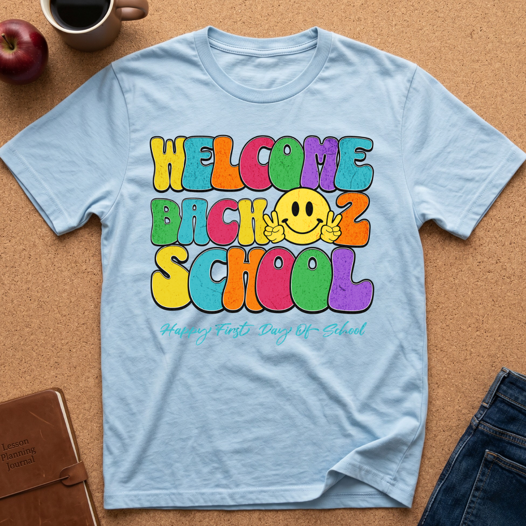 A light blue t-shirt laid flat on a cork background, featuring a colorful retro-style graphic that reads 'WELCOME BACK 2 SCHOOL' with a yellow smiley face making peace signs. Styled flat-lay props include an apple, coffee mug, notebook, and jeans.