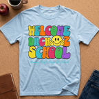 A light blue t-shirt laid flat on a cork background, featuring a colorful retro-style graphic that reads 'WELCOME BACK 2 SCHOOL' with a yellow smiley face making peace signs. Styled flat-lay props include an apple, coffee mug, notebook, and jeans.