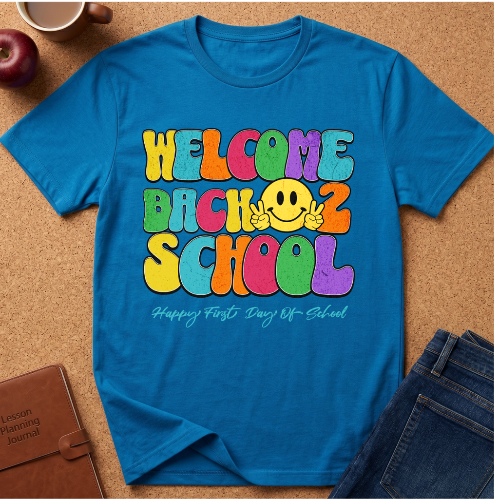 A bright blue graphic t-shirt laid flat on a cork background, featuring a colorful retro-style 'WELCOME BACK 2 SCHOOL' design with a yellow smiley face and the phrase 'Happy First Day Of School' underneath. Styled flat-lay props include a brown leather journal, an apple, a coffee cup, and blue jeans.