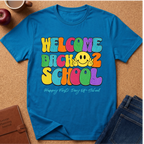 A bright blue graphic t-shirt laid flat on a cork background, featuring a colorful retro-style 'WELCOME BACK 2 SCHOOL' design with a yellow smiley face and the phrase 'Happy First Day Of School' underneath. Styled flat-lay props include a brown leather journal, an apple, a coffee cup, and blue jeans.