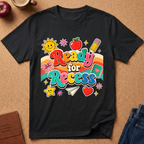 A black graphic t-shirt laid flat on a cork background, featuring a colorful retro-style 'Ready for Recess' design with school-themed illustrations like a sun, apple, pencil, and stars.