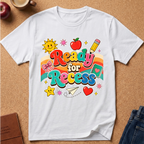 A white short-sleeve t-shirt with a colorful 'Ready for Recess' graphic print, laid flat on a cork background with school-themed props like an apple, a pencil, and a star.