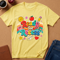 A yellow short-sleeve t-shirt with a colorful 'Ready for Recess' graphic, laid flat on a cork background with lifestyle props including a coffee mug, an apple, a notebook, and blue jeans.