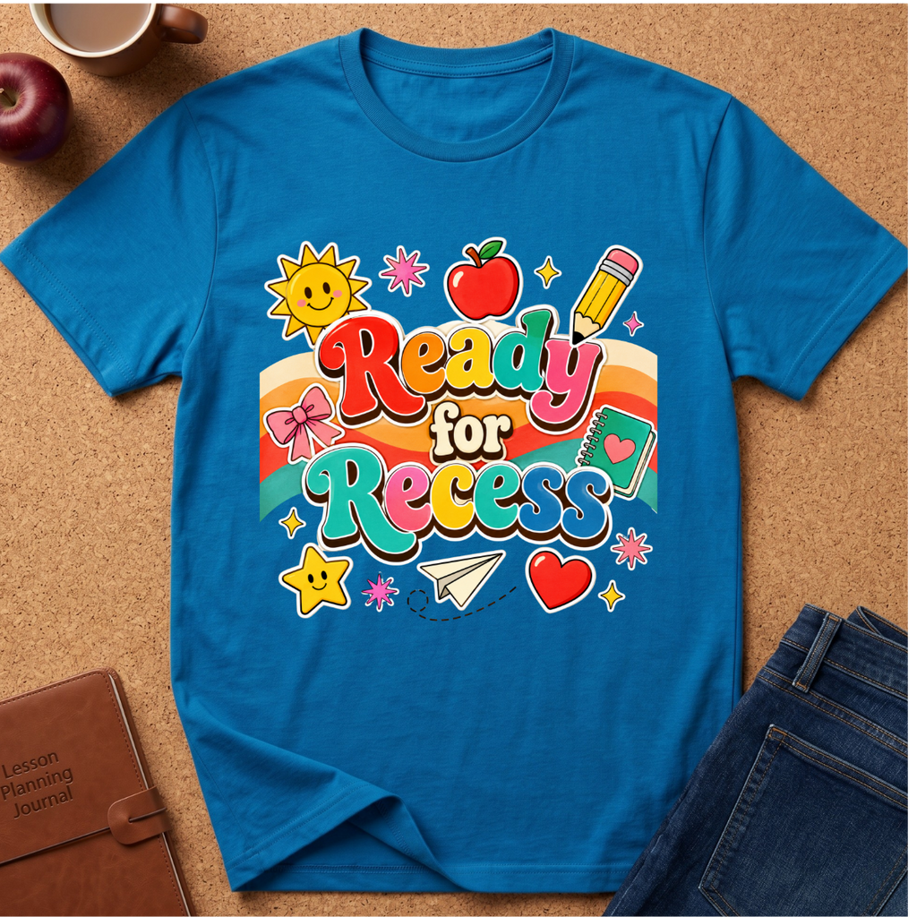 A bright blue short-sleeve t-shirt with a colorful retro 'Ready for Recess' graphic, styled in a flat-lay on a cork background with an apple, coffee cup, notebook, and jeans.