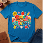 A bright blue short-sleeve t-shirt with a colorful retro 'Ready for Recess' graphic, styled in a flat-lay on a cork background with an apple, coffee cup, notebook, and jeans.
