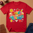 A red short-sleeve graphic T-shirt laid flat on a cork surface, featuring colorful retro-style text reading 'Ready for Recess' with school-themed illustrations including a sun, apple, pencil, bow tie, notebook, stars, and a heart. Partial props around the shirt include an apple, a cup, a notebook, and jeans.