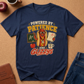 A navy blue graphic t-shirt laid flat on a brown cork background, surrounded by lifestyle props including a leather journal, an apple, a coffee cup, and the corner of a pair of jeans. The t-shirt features a colorful cartoon graphic of a hot dog character wearing glasses and holding a paper with an A+, next to a chalkboard and apple, with the text 'POWERED BY PATIENCE AND GLIZZYS'.