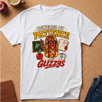 A white short-sleeve graphic T-shirt laid flat on a cork surface, surrounded by small styled props including an apple, a mug, a notebook, and folded jeans. The shirt features a large colorful front print with the text 'POWERED BY PATIENCE AND GLIZZYS' and an illustrated hot-dog character wearing glasses and holding a grade A paper.