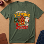 An olive green graphic t-shirt laid flat on a cork background, featuring a cartoon hot dog character with glasses and the text 'POWERED BY PATIENCE AND GLIZZYS'.