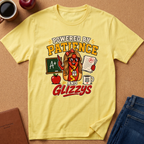 A yellow graphic t-shirt laid flat on a cork surface, surrounded by lifestyle props including a coffee mug, an apple, a notebook, and folded blue jeans. The t-shirt features a colorful cartoon graphic of a hot dog character wearing glasses and a lanyard, with the text 'POWERED BY PATIENCE AND GLIZZYS'.