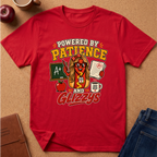 A red short-sleeve graphic T-shirt laid flat on a tan cork-like surface with school-themed props around it, including an apple, mug, notebook, and jeans. The shirt features a large colorful front print reading “POWERED BY PATIENCE AND GLIZZYS” with an illustrated hot dog character, chalkboard, apple, coffee mug, and papers.