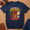 A navy blue graphic t-shirt laid flat on a brown cork background, surrounded by lifestyle props including an apple, a coffee cup, a leather journal, and the corner of blue jeans. The t-shirt features a colorful cartoon graphic of a hot dog character wearing glasses and a teacher badge, with the text 'POWERED BY PATIENCE AND GLZZYS'.