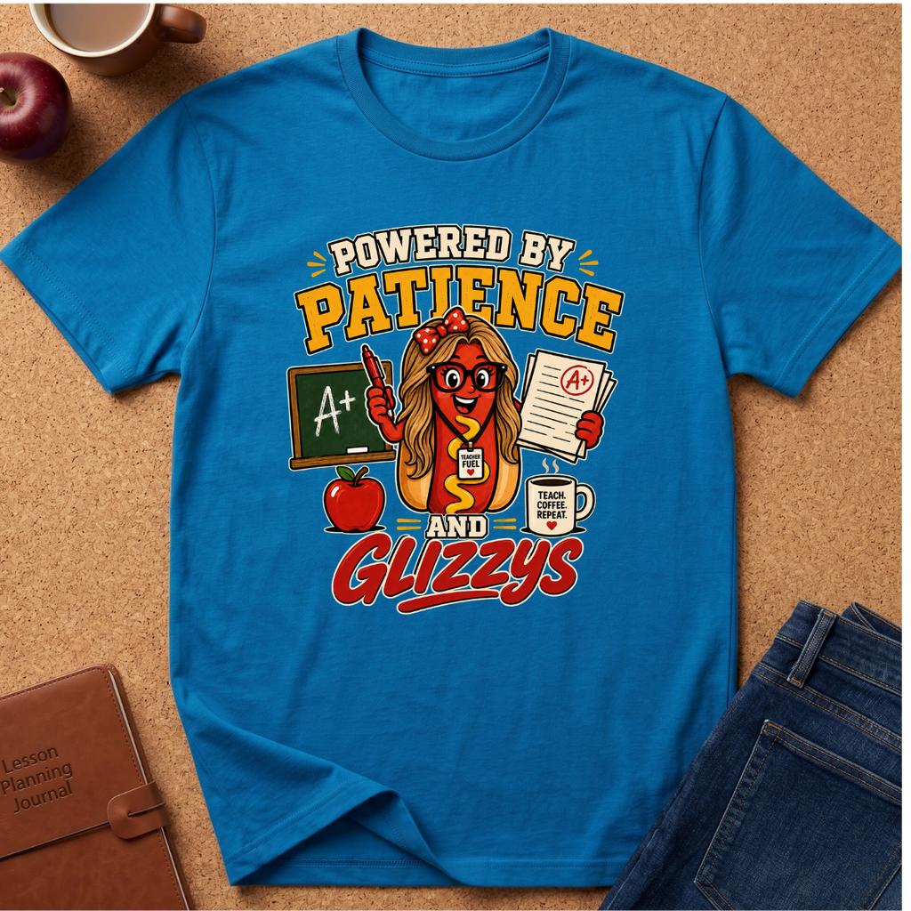 A bright blue graphic t-shirt laid flat on a cork background, featuring a cartoon hot dog character with glasses and the text 'POWERED BY PATIENCE AND GLIZZYS' along with school-themed icons like an apple, chalkboard, and coffee mug. Styled flat-lay props include a notebook, apple, coffee cup, and jeans.